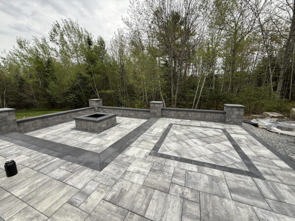 Large gray paver patio with a square fire pit and low seat wall bordered by charcoal accent stones.
