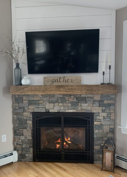 Stone-surround gas fireplace below a wall-mounted TV with a wood mantel and gather sign