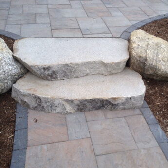 Rough-cut granite steps flanked by round boulders on a paver patio