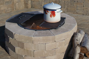 Round tan concrete block fire pit kit with a cooking grate and enamel pot on top.
