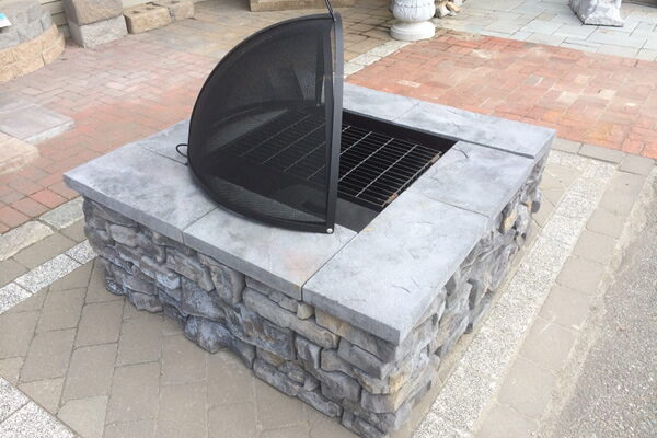 Square gray stone fire pit with mesh spark screen open on a brick paver patio