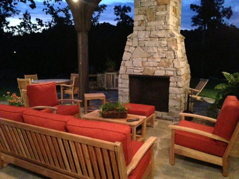 Teak outdoor sectional sofa and chairs with red cushions around a stone fireplace at dusk
