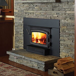 Black wood-burning fireplace insert with an arched door in a stacked stone surround, firewood nearby.