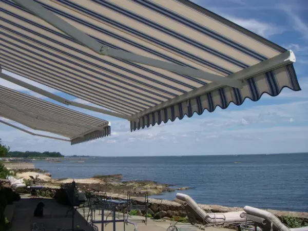 Striped retractable awnings over an oceanfront patio with furniture and a rocky shoreline.