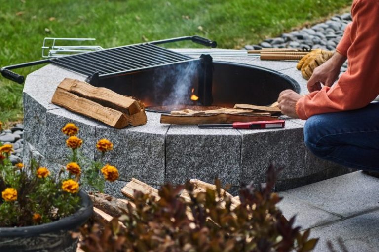 Person lighting a round granite block fire pit with a swing-away cooking grate and firewood.