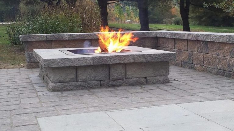 Square stone block gas fire pit burning on a patio beside a seat wall