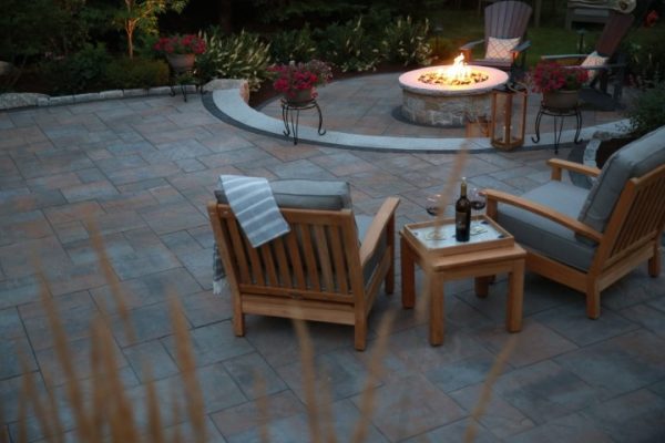 Paver patio with a lit stone fire pit, teak lounge chairs, and flowering potted plants at dusk