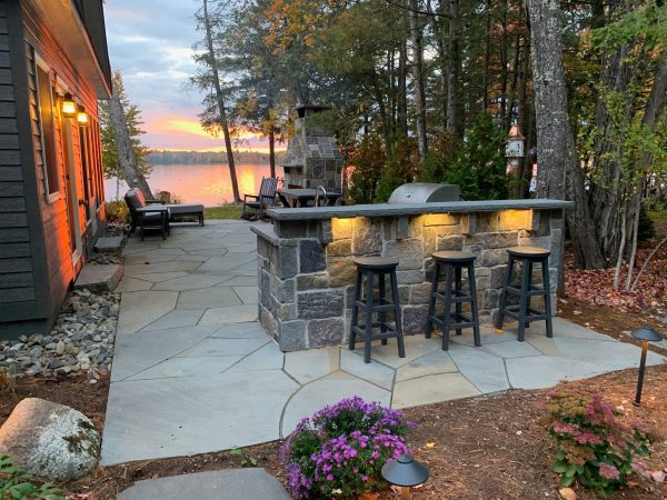 Stone outdoor bar with integrated lighting and stools on a patio overlooking a lake at sunset.