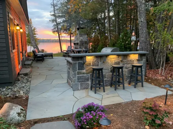 Stone outdoor kitchen bar with a grill and three stools lit by path lighting, overlooking a lake at sunset.