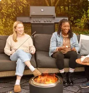 Two women roasting marshmallows at a round steel fire pit beside a gas grill on a patio.