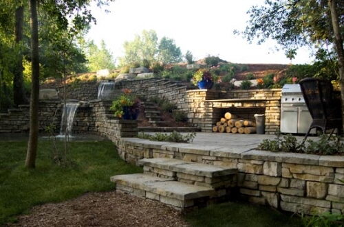 Tiered stone retaining wall patio with a waterfall, fireplace, and grill on a landscaped slope.
